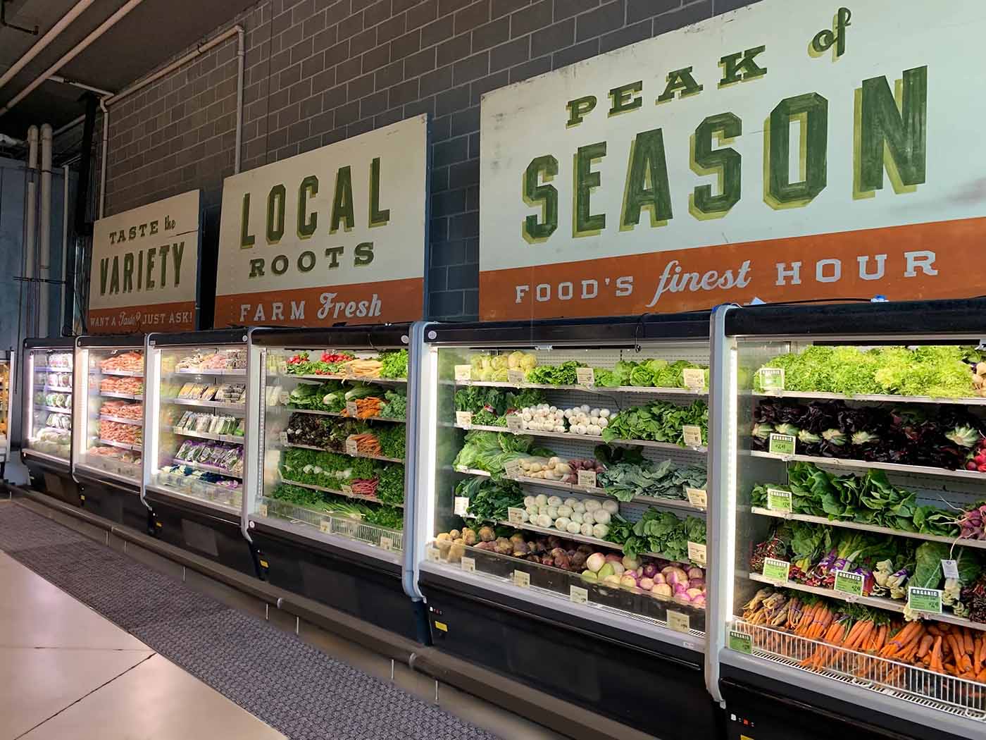 Refrigerated display showcasing fresh produce, with green and orange signage promoting local and seasonal foods.