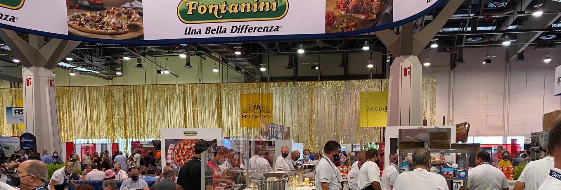 Busy exhibition hall featuring a gold backdrop and attendees engaging with food displays.