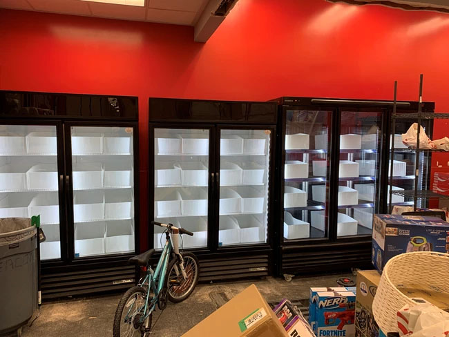 Empty black refrigerator units against a red wall, with a bicycle and cardboard boxes in the foreground.