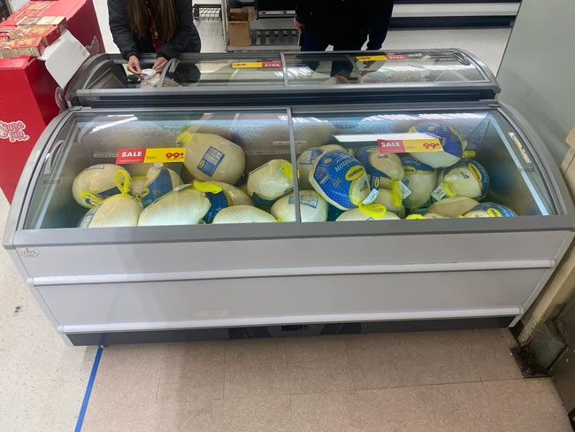 Frozen turkeys in a display case, featuring blue and yellow packaging, with shoppers nearby.